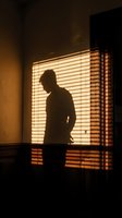 Silhouette of male figure standing against warm glowing window with horizontal blinds casting striped shadows on wall, creating dramatic lighting effect for interior design and mood concepts.