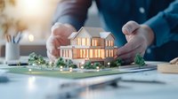 Illuminated architectural model of luxury house held by hands in professional setting. Scale model shows modern residential design with landscaping and warm interior lighting.