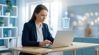 Woman in business casual attire working on laptop at wooden desk in modern office with blue shelving and natural light for corporate, professional, and workplace concepts.