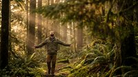 Young Caucasian man in camouflage jacket with arms outstretched walking through misty forest path surrounded by tall trees and ferns during golden hour sunlight.
