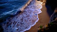 Gentle ocean waves washing up on sandy beach at sunset, creating white foam patterns along shoreline with dramatic lighting and deep blue water contrasts.
