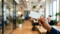 Young professional man of African descent holding blank white business card in modern office environment with blurred colleagues and plants in background, suitable for corporate branding and business networking materials.