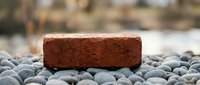 Red clay brick resting on smooth gray pebbles with soft blurred natural background, symbolizing construction, strength, and building materials.