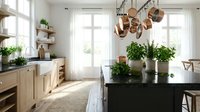 Modern farmhouse kitchen interior with natural wood cabinets, black countertops, hanging copper pots, white subway tiles, and fresh green plants by sunny windows.