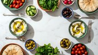 Mediterranean breakfast spread with hummus, olives, fresh vegetables, pita bread and herbs on white wooden table. Healthy Middle Eastern cuisine.