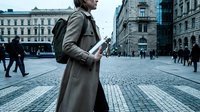Young professional woman walking through historic city square while using smartphone during commute in urban European setting with cobblestone streets.
