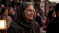 Elderly Middle Eastern woman wearing traditional headscarf smiling warmly at cultural event with soft lighting and blurred crowd background.
