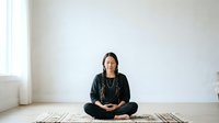 Mature woman of South Asian descent sitting in lotus position on wooden floor in minimalist room, wearing black clothing and glasses, meditating or practicing yoga for wellness and mindfulness content.