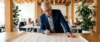 Senior male architect with gray hair and glasses reviewing blueprints at wooden desk in modern office with exposed beams, potted plants, and colleagues working in background.