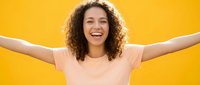 Happy young woman with curly brown hair and medium skin tone wearing peach t-shirt, arms outstretched against bright yellow background, expressing joy and freedom.