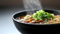 Steaming bowl of Asian noodle soup with tender sliced meat, green onions and savory broth, served in traditional black ceramic bowl against dark background.