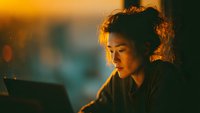 Young Asian woman working late on laptop with warm golden lighting creating focused atmosphere for productivity and remote work concepts.