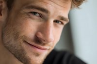 Handsome young Caucasian man with stubble and wavy hair smiling confidently at camera in close-up portrait for lifestyle and business content.