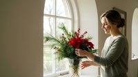 Young woman arranging festive red flowers and evergreen branches in glass vase by arched window in bright interior for holiday home decoration.