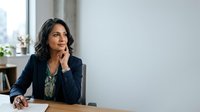 Thoughtful woman professional in dark blazer sitting at desk with hand near chin, gazing upward in contemplative pose, suitable for business, leadership, and corporate concepts.