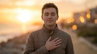 Young man with hand on chest at sunset, expressing gratitude, sincerity, or pledge against urban waterfront backdrop with golden hour lighting.