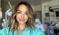 Young female medical professional in turquoise scrubs takes selfie with confident smile and wavy blonde hair, hospital operating room and staff visible in background.
