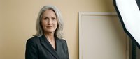 Mature woman with long gray hair wearing dark blazer in modern office setting with neutral beige walls and contemporary decor.