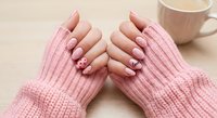 Female hands with pink manicured nails in cozy knit sweater sleeves holding warm coffee mug on wooden table creating hygge atmosphere for relaxation.