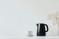 Minimalist kitchen scene with black electric kettle and white coffee cup on light surface against neutral wall, dried flowers in vase add natural accent.
