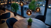 Modern dining area with round glass pendant lights casting warm glow over marble table, navy velvet chairs and potted plant, evening city view through windows.