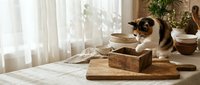 Siamese cat exploring wooden kitchen utensils and cutting board on sunlit table with natural home decor and soft window lighting.