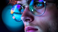 Young caucasian man wearing glasses illuminated by vibrant neon lights creating atmospheric bokeh effect, close up portrait with focus on bright blue eye and reflective lens.