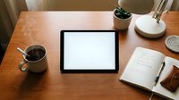 Digital tablet with blank white screen on wooden desk workspace with coffee mug, notebook, pen and small plant for productivity and creative work mockup.