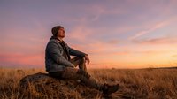 Middle-aged Caucasian male farmer sitting on log in golden wheat field at sunset, contemplating rural landscape. Agricultural lifestyle and harvest season concept.