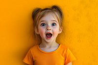 Excited young girl with wide open mouth and surprised expression wearing orange t-shirt against vibrant yellow wall background showing pure joy and wonder.