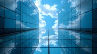 Modern glass office buildings viewed from below against blue sky with white clouds reflecting in windows, creating symmetrical architectural perspective.