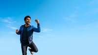 Man in casual athletic wear jumping with fists raised against clear blue sky, expressing joy and success for motivational, lifestyle, and wellness content.