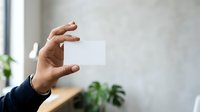 Hand holding blank white card in modern office space with plants and natural light for branding and marketing presentations.