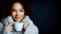 Young African American woman with curly hair smiling while holding white coffee mug, wearing cozy knit sweater against dark background, expressing warmth and contentment.