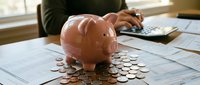 Pink piggy bank surrounded by coins on financial documents with person using calculator in background for budgeting and savings planning.