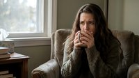 Woman with long dark hair and fair complexion sitting in armchair, wearing beige knit sweater, hand near mouth showing worried or anxious expression, natural window light, indoor home setting for mental health, stress, concern themes.