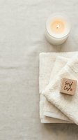 Soft folded towels with a glowing candle and wooden self care block on linen, evoking calm wellness and spa relaxation routines.