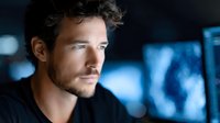 Young Caucasian male professional with curly hair in dark clothing against blurred blue technology background, contemplating future innovations in modern office.