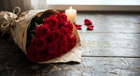 Red roses bouquet wrapped in kraft paper with burning candle on rustic wooden table creating romantic atmosphere for Valentine's Day celebration.