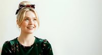 Young woman with blonde hair, fair complexion, wearing dark green sequined top and sunglasses on head, smiling at camera against white background for portrait and lifestyle use.