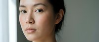 Young Asian woman with natural makeup looking thoughtfully at camera in soft natural light. Professional headshot for wellness and lifestyle content.