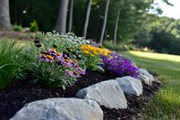 Colorful flower garden bed bordered with large gray rocks and fresh mulch, featuring purple, yellow and pink blooms against lush green foliage in summer landscape.