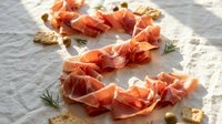 Sliced prosciutto ham with rosemary herbs and nuts arranged on white parchment paper for gourmet appetizer and charcuterie board presentation.