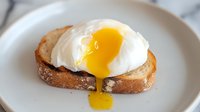 Poached egg on toasted bread slice with bright yellow yolk dripping down, served on white ceramic plate. Close up view of breakfast preparation.