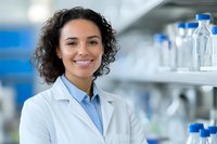 Hispanic female scientist in lab coat smiling confidently in modern laboratory setting with research equipment and glass containers on shelves in background.