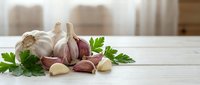 Fresh garlic bulbs and cloves with green parsley leaves arranged on white wooden table in bright kitchen interior for cooking and culinary preparations.
