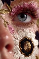 Close-up of eye with artistic makeup featuring pink eyeshadow, gold accents, and intricate embroidery-like floral design on face for high fashion or theatrical use.