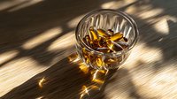 Glass of whiskey with ice cubes on wooden table in dramatic sunlight with shadows and light reflections creating atmospheric mood.