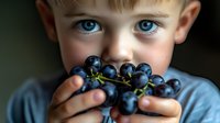 Young boy with bright blue eyes holding fresh dark purple grapes close to face, creating playful and healthy eating concept in moody natural lighting.