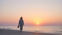 Woman in casual clothing walking along sandy beach toward golden sunset over calm ocean waters, suitable for travel, lifestyle, and wellness marketing materials.